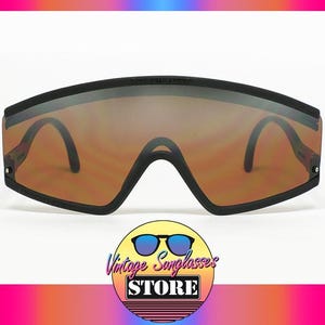 Op de afbeelding: Zwarte vintage zonnebril met bruin getinte glazen. De zonnebril heeft een breed, rechthoekig montuur en een brug die naar beneden buigt. De pootjes zijn dun en zwart. Het logo van Vintage Sunglasses Store staat onderaan.