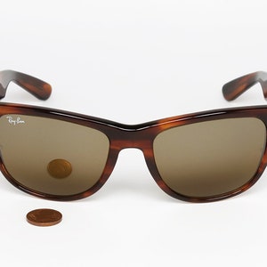 Peut inclure: Lunettes de soleil Ray-Ban Wayfarer en &eacute;caille de tortue avec des verres bruns. Les lunettes de soleil sont pos&eacute;es sur une surface blanche avec un penny pour l'&eacute;chelle.