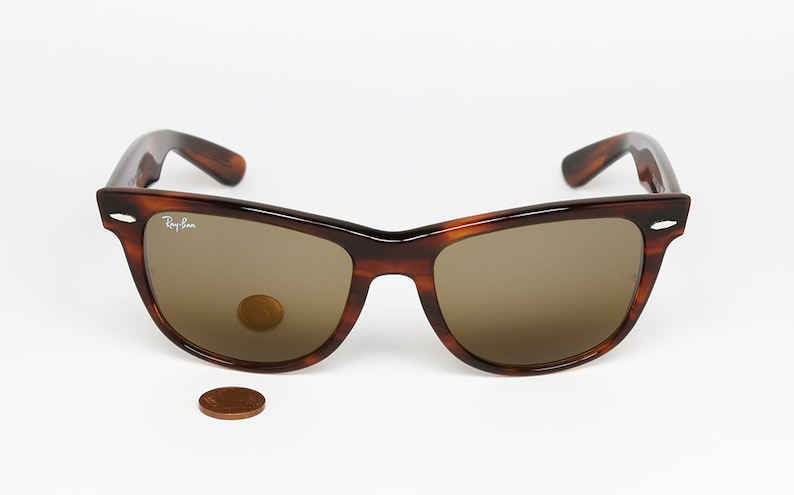 Peut inclure: Lunettes de soleil Ray-Ban Wayfarer en &eacute;caille de tortue avec des verres bruns. Les lunettes de soleil sont pos&eacute;es sur une surface blanche avec un penny pour l'&eacute;chelle.
