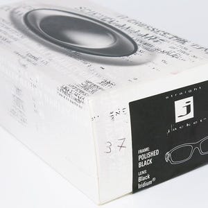 May include: A white and black box for Straight Jacket sunglasses. The box features a black label with the brand name and a drawing of the glasses. The frame is polished black with black iridium lenses. The number 37 is written on the box.