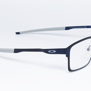 K&ouml;nnte beinhalten: Rechteckige Brille in Schwarz und Grau mit klaren Gl&auml;sern. Die Fassungen sind dunkelmarineblau mit grauen B&uuml;geln. Das Oakley-Logo ist auf dem B&uuml;gel sichtbar. Die Brille ist vor einem wei&szlig;en Hintergrund platziert.