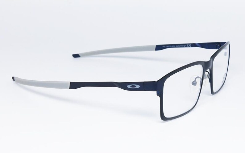 K&ouml;nnte beinhalten: Rechteckige Brille in Schwarz und Grau mit klaren Gl&auml;sern. Die Fassungen sind dunkelmarineblau mit grauen B&uuml;geln. Das Oakley-Logo ist auf dem B&uuml;gel sichtbar. Die Brille ist vor einem wei&szlig;en Hintergrund platziert.