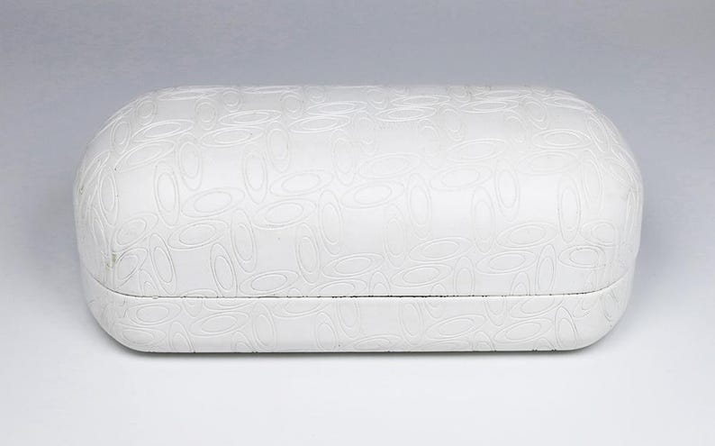 Puede incluir: Un estuche blanco ovalado para gafas con una superficie texturizada que presenta un patr&oacute;n repetitivo de formas ovaladas. El estuche cerrado parece estar hecho de un material duradero, adecuado para proteger las gafas.
