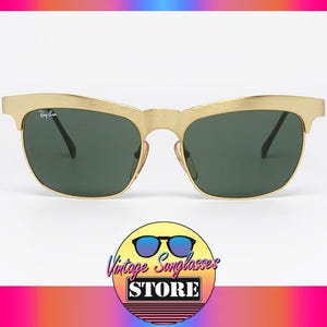 Puede incluir: Gafas de sol doradas con lentes verde oscuro. El logotipo de Ray-Ban es visible en la lente izquierda. Las gafas de sol tienen un dise&ntilde;o retro. La imagen tambi&eacute;n incluye un logotipo de una tienda de gafas de sol vintage.