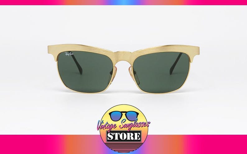 Puede incluir: Gafas de sol doradas con lentes verde oscuro. El logotipo de Ray-Ban es visible en la lente izquierda. Las gafas de sol tienen un dise&ntilde;o retro. La imagen tambi&eacute;n incluye un logotipo de una tienda de gafas de sol vintage.