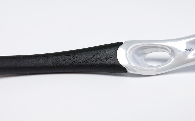 Pu&ograve; includere: Primo piano di una parte di occhiali sportivi. L'asta nera presenta la scritta "Radar" in corsivo. L'altra estremit&agrave; &egrave; argentata con un ritaglio ovale e il numero "09-673".