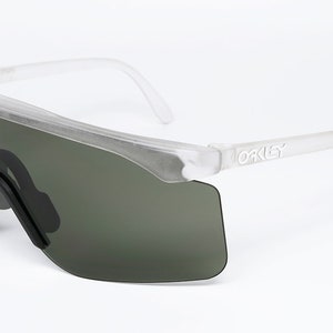 Puede incluir: Gafas de sol con montura transparente y lentes verde oscuro con la palabra "Oakley" impresa en el lateral.