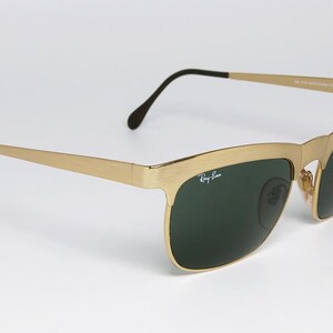 Puede incluir: Gafas de sol doradas con lentes verde oscuro. Las gafas tienen una montura rectangular y patillas negras. El logotipo de Ray-Ban es visible en la lente izquierda. Las gafas est&aacute;n sobre un fondo blanco.