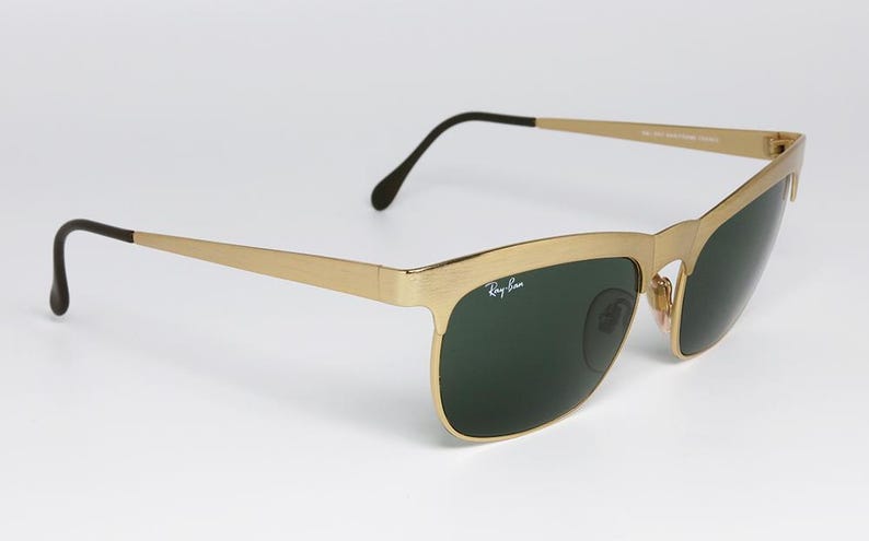 Puede incluir: Gafas de sol doradas con lentes verde oscuro. Las gafas tienen una montura rectangular y patillas negras. El logotipo de Ray-Ban es visible en la lente izquierda. Las gafas est&aacute;n sobre un fondo blanco.