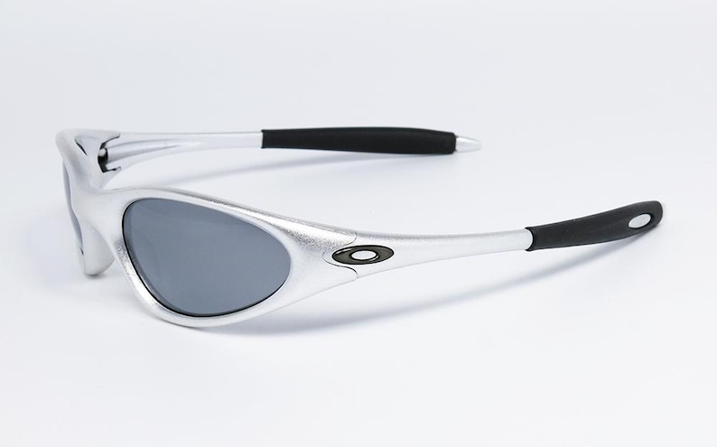 May include: Silver-framed sunglasses with gray lenses. The sunglasses have black temple tips and a small logo on the temples. The design is sleek and modern, suitable for outdoor activities.