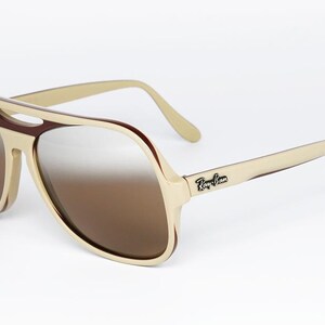 May include: Cream-colored aviator sunglasses with brown gradient lenses. The frames have a retro design with a brown accent around the lenses and the Ray-Ban logo on the temple.