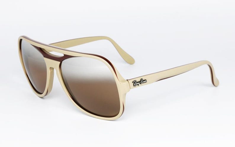 May include: Cream-colored aviator sunglasses with brown gradient lenses. The frames have a retro design with a brown accent around the lenses and the Ray-Ban logo on the temple.