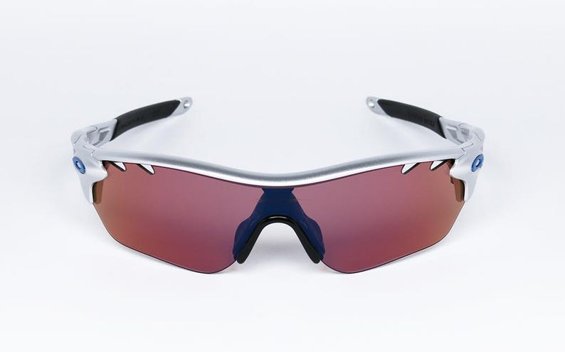 May include: Silver framed sunglasses with red tinted lenses. The sunglasses have a sporty design with a curved frame.