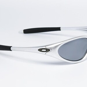 May include: Silver-framed sunglasses with gray lenses. The sunglasses have black accents on the temples and a small logo on the frame. The sleek design is suitable for outdoor activities.
