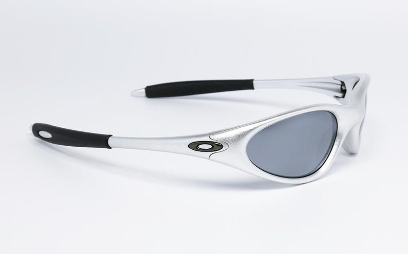 May include: Silver-framed sunglasses with gray lenses. The sunglasses have black accents on the temples and a small logo on the frame. The sleek design is suitable for outdoor activities.