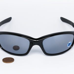 May include: Black sunglasses with gray lenses, featuring a curved design and a small blue and white sticker on the right lens. A penny is placed below the sunglasses. The sunglasses are on a white background.