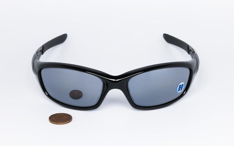 May include: Black sunglasses with gray lenses, featuring a curved design and a small blue and white sticker on the right lens. A penny is placed below the sunglasses. The sunglasses are on a white background.