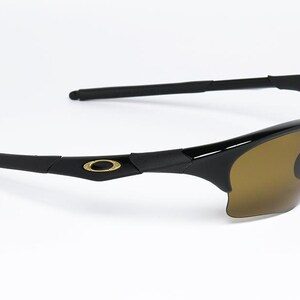 May include: Black sunglasses with brown tinted lenses. The sunglasses have a sleek, modern design with a gold emblem on the temples. The arms are straight and extend back from the frame.