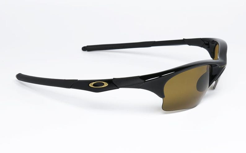 May include: Black sunglasses with brown tinted lenses. The sunglasses have a sleek, modern design with a gold emblem on the temples. The arms are straight and extend back from the frame.