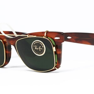 May include: Tortoiseshell-framed sunglasses with a clip-on design. The sunglasses feature dark green lenses and gold-colored metal accents. The Ray-Ban logo is visible on the lens. The glasses have a classic, retro style.