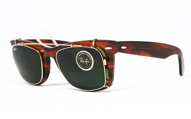 May include: Tortoiseshell-framed sunglasses with a clip-on design. The sunglasses feature dark green lenses and gold-colored metal accents. The Ray-Ban logo is visible on the lens. The glasses have a classic, retro style.