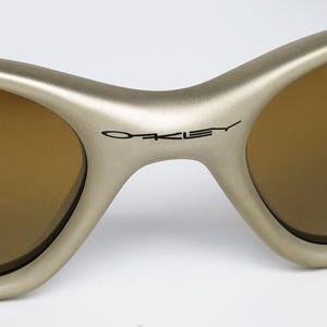 May include: Close-up of Oakley sunglasses with a matte gold frame and brown lenses. The Oakley logo is printed in black on the bridge. The sunglasses have a curved, sporty design.