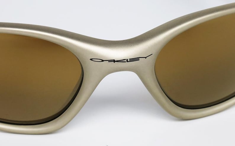 May include: Close-up of Oakley sunglasses with a matte gold frame and brown lenses. The Oakley logo is printed in black on the bridge. The sunglasses have a curved, sporty design.
