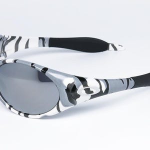 May include: Gray and white camouflage sunglasses with dark gray lenses. The arms of the sunglasses are black at the ends. The sunglasses are set against a white background.