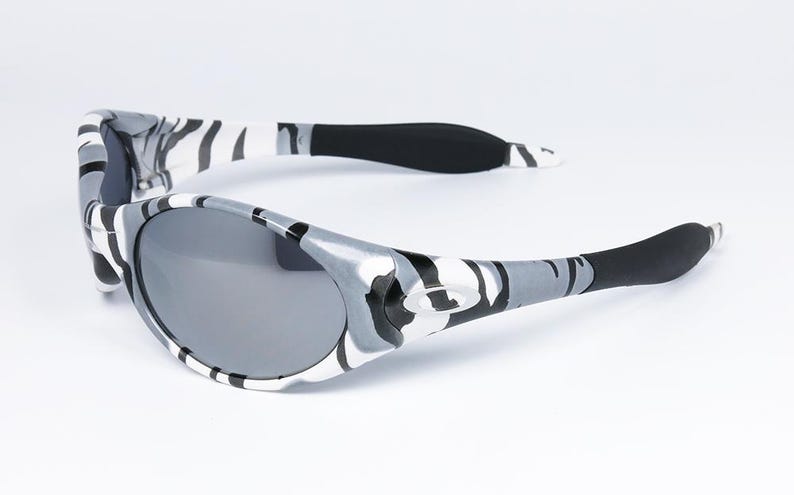 May include: Gray and white camouflage sunglasses with dark gray lenses. The arms of the sunglasses are black at the ends. The sunglasses are set against a white background.
