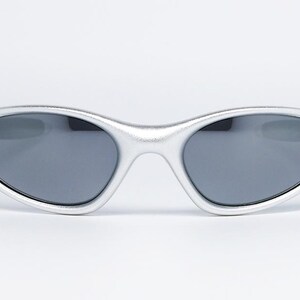 May include: Silver-framed sunglasses with dark gray lenses. The oval-shaped frames have a metallic finish. The sunglasses are designed with a sleek, modern aesthetic, suitable for various outdoor activities.