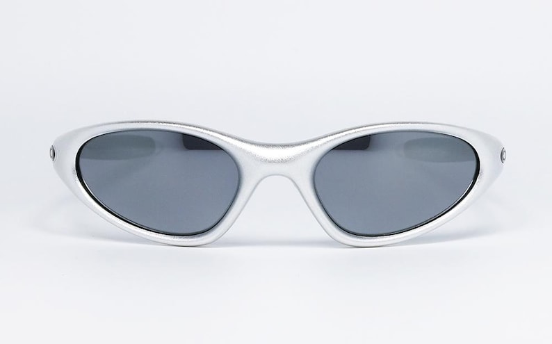 May include: Silver-framed sunglasses with dark gray lenses. The oval-shaped frames have a metallic finish. The sunglasses are designed with a sleek, modern aesthetic, suitable for various outdoor activities.