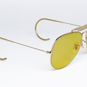 K&ouml;nnte beinhalten: Goldene Aviator-Sonnenbrille mit gelben Gl&auml;sern. Die Sonnenbrille hat ein klassisches Design mit einem d&uuml;nnen Metallrahmen und Drahtb&uuml;geln.