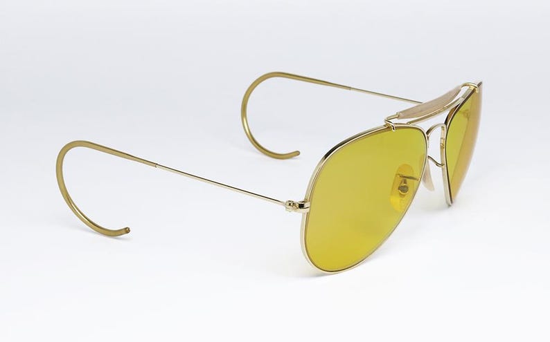 K&ouml;nnte beinhalten: Goldene Aviator-Sonnenbrille mit gelben Gl&auml;sern. Die Sonnenbrille hat ein klassisches Design mit einem d&uuml;nnen Metallrahmen und Drahtb&uuml;geln.