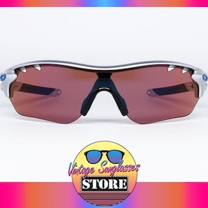 May include: Silver framed sunglasses with brown lenses. The sunglasses have a sporty design with a wraparound frame.