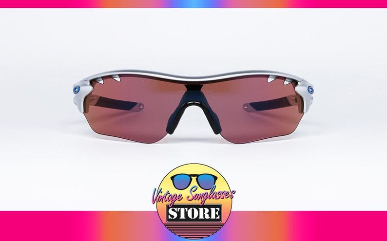 May include: Silver framed sunglasses with brown lenses. The sunglasses have a sporty design with a wraparound frame.