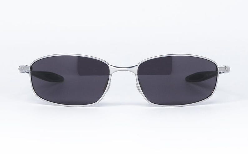 May include: Silver-framed sunglasses with dark gray lenses. The sunglasses have a rectangular shape with rounded edges. The frame is thin and metallic, with hinges on the sides. The lenses are a solid, dark gray color, providing a sleek and modern look.