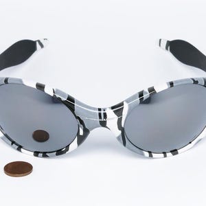 May include: Gray and white camouflage sunglasses with dark gray lenses. The sunglasses have a rounded shape and black ear pieces. A penny is next to the sunglasses for size comparison.