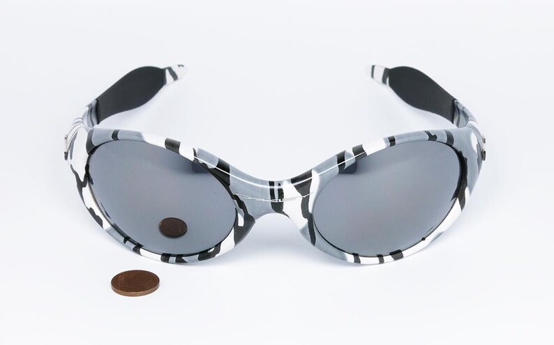 May include: Gray and white camouflage sunglasses with dark gray lenses. The sunglasses have a rounded shape and black ear pieces. A penny is next to the sunglasses for size comparison.