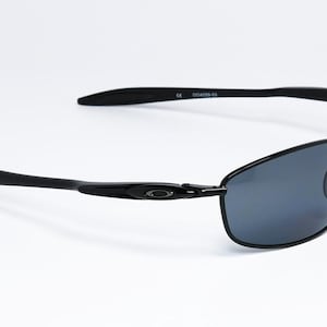 Puede incluir: Gafas de sol rectangulares negras con lentes grises oscuras. Las gafas tienen un dise&ntilde;o elegante y moderno con montura y patillas negras. El logotipo de Oakley es visible en la patilla. Las gafas est&aacute;n sobre un fondo blanco.