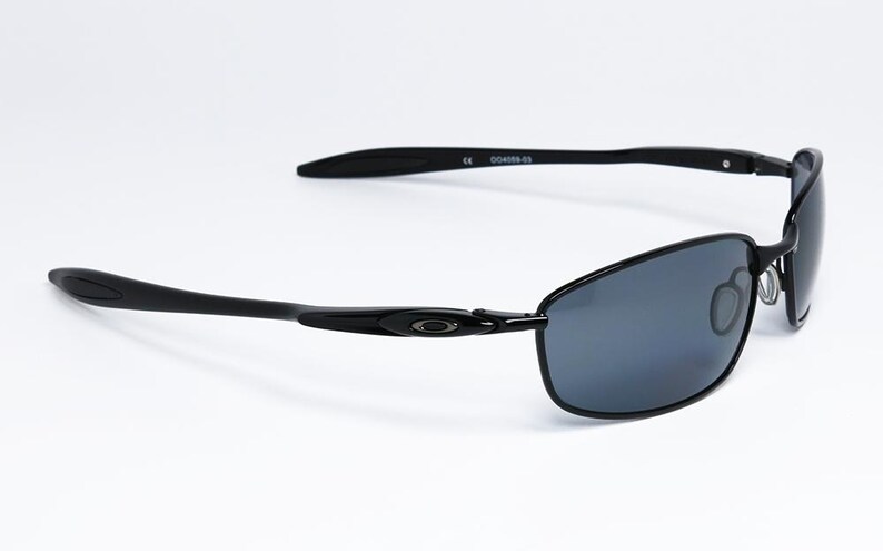 Puede incluir: Gafas de sol rectangulares negras con lentes grises oscuras. Las gafas tienen un dise&ntilde;o elegante y moderno con montura y patillas negras. El logotipo de Oakley es visible en la patilla. Las gafas est&aacute;n sobre un fondo blanco.