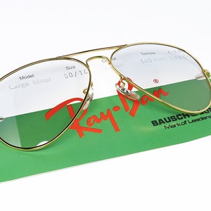 Op de afbeelding: Gouden Ray-Ban aviator zonnebril met heldere glazen. De zonnebril ligt op een groen-witte kaart met de tekst "Ray-Ban" en "Bausch & Lomb Mark of Leadership".