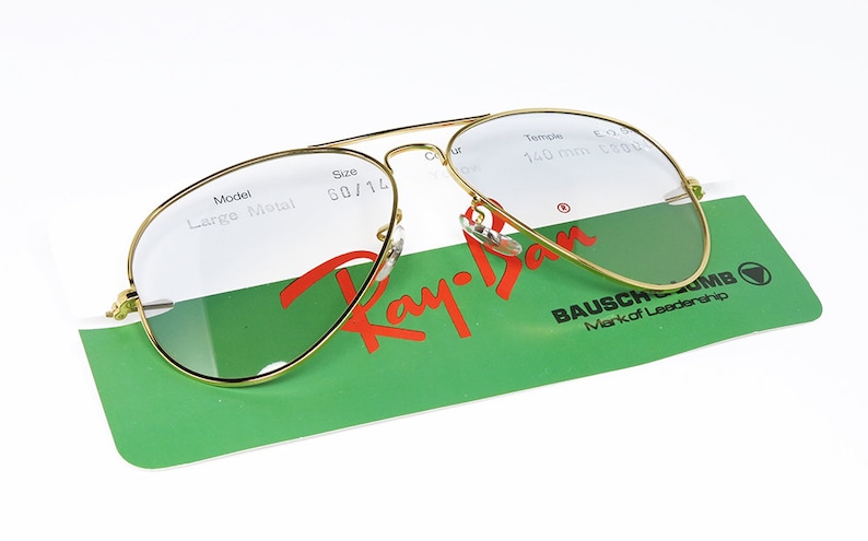 Op de afbeelding: Gouden Ray-Ban aviator zonnebril met heldere glazen. De zonnebril ligt op een groen-witte kaart met de tekst "Ray-Ban" en "Bausch & Lomb Mark of Leadership".