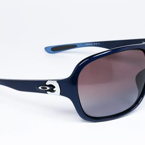 May include: Dark navy blue sunglasses with a wraparound design. The sunglasses have a glossy finish, brown gradient lenses, and the Oakley logo on the temples. The arms are a lighter shade of blue at the tips.