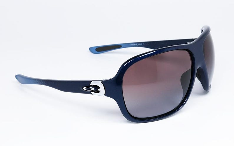 May include: Dark navy blue sunglasses with a wraparound design. The sunglasses have a glossy finish, brown gradient lenses, and the Oakley logo on the temples. The arms are a lighter shade of blue at the tips.