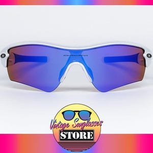 Pu&ograve; includere: Occhiali da sole con montatura bianca e lenti blu e viola. Il logo Oakley &egrave; visibile sulle lenti. L'immagine include un logo per Vintage Sunglasses Store.
