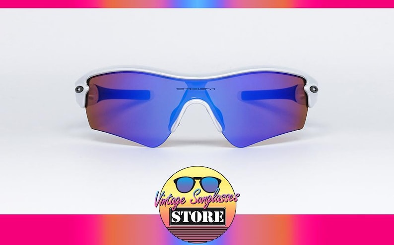 Pu&ograve; includere: Occhiali da sole con montatura bianca e lenti blu e viola. Il logo Oakley &egrave; visibile sulle lenti. L'immagine include un logo per Vintage Sunglasses Store.