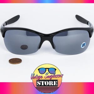 May include: Black sunglasses with gray lenses. The sunglasses have a curved frame and a small logo on the lens. A penny is next to the sunglasses. The logo for Vintage Sunglasses Store is in the bottom right corner.