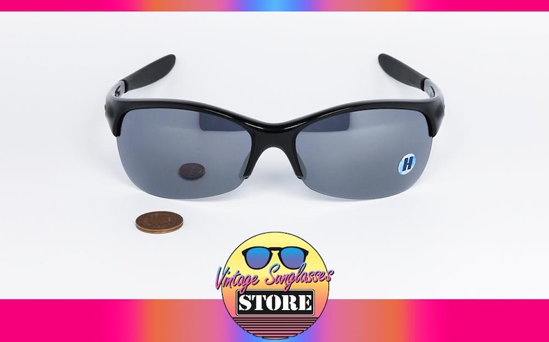 May include: Black sunglasses with gray lenses. The sunglasses have a curved frame and a small logo on the lens. A penny is next to the sunglasses. The logo for Vintage Sunglasses Store is in the bottom right corner.