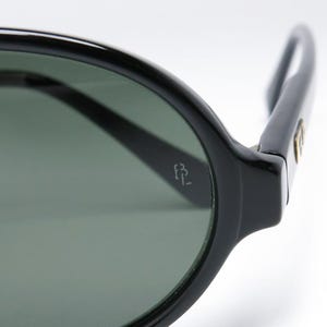 May include: Close-up of black sunglasses with dark green lenses. The frame is thick and rounded, with a small logo on the temple. The sunglasses are set against a white background.