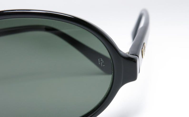 May include: Close-up of black sunglasses with dark green lenses. The frame is thick and rounded, with a small logo on the temple. The sunglasses are set against a white background.